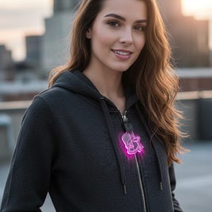 Könnte beinhalten: Dunkelgrauer Kapuzenpullover mit Reißverschluss und einem neonrosa Anhänger. Der Anhänger hat eine leuchtende Kontur. Der Hoodie hat eine Kapuze mit Kordelzug und Vordertaschen. Im Hintergrund eine Stadtlandschaft.