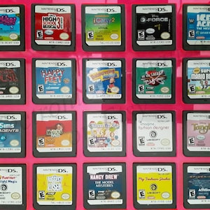 Puede incluir: Una colección de 18 cartuchos de juegos de Nintendo DS que se muestran en una caja de plástico rosa. Los cartuchos están dispuestos en tres filas de seis. Los juegos incluyen títulos como "Monster House", "Happy Feet", "My Sims Agents", "Word World", "Cheer We Go", "Tangled", "Nancy Drew: The Model Mysteries" y "My Fashion Studio".