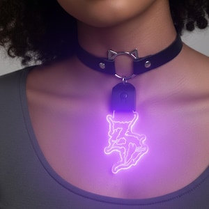 Puede incluir: Collar gargantilla de cuero negro con diseño de orejas de gato plateadas. Un colgante brillante púrpura cuelga de la gargantilla, con un diseño estilizado. El colgante está iluminado con una luz púrpura brillante.