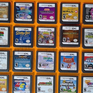 Könnte beinhalten: Eine Sammlung von 20 Nintendo DS-Spielkassetten, die in einem durchsichtigen Plastik-Etui mit gelbem Rahmen ausgestellt sind. Die Kassetten sind in vier Reihen zu fünf angeordnet. Die Kassetten sind alle in verschiedenen Farben und haben unterschiedliche Titel.