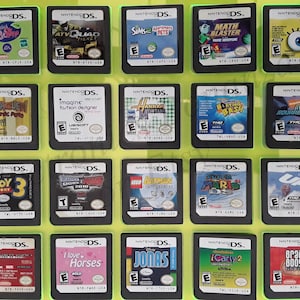 May include: A collection of 18 Nintendo DS game cartridges displayed in a clear plastic case. The cartridges are arranged in a grid of three rows and six columns. The cartridges are all different colors and have different game titles printed on them.