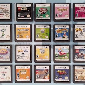 May include: A collection of 16 Nintendo DS game cartridges displayed in a clear plastic case. The cartridges are arranged in four rows of four, with each cartridge featuring a different game title and cover art. Some of the games include "LEGO Star Wars", "Guitar Hero", "The Sims", "Tangled", and "Dora the Explorer".