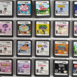 May include: A collection of 18 Nintendo DS game cartridges, displayed in a grid on a white surface. The cartridges feature a variety of popular children's characters and themes, including Dora the Explorer, Hello Kitty, Plants vs. Zombies, and Cooking Mama.