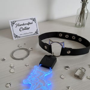 Puede incluir: Collar de cuero negro con un centro plateado en forma de oreja de gato. Un colgante azul brillante cuelga del collar. La imagen también incluye una cadena de plata, abalorios y un pequeño candado. Una tarjeta dice "Handcrafted Collar".