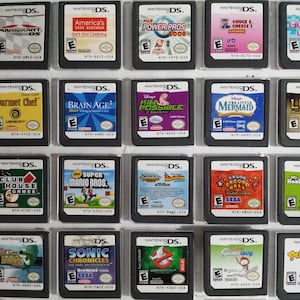 Op de afbeelding: Een verzameling van 18 Nintendo DS-spelcartridges, tentoongesteld in een doorzichtige plastic koffer. De cartridges zijn gerangschikt in drie rijen van zes, waarbij elke cartridge een andere speltitel en coverart heeft. Sommige van de spellen zijn Mario Kart DS, Brain Age 2 en De Kleine Zeemeermin.