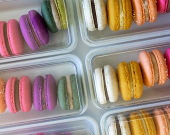 May include: A collection of colorful macarons arranged in clear plastic rectangular trays. The macarons feature various shades including yellow, orange, pink, purple, and green, each with a visible cream filling sandwiched between two round cookies.