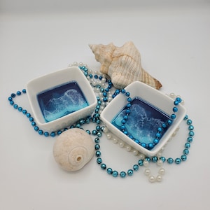 May include: Two white ceramic dishes with blue resin ocean wave designs. The dishes are surrounded by a string of white and blue beads and a seashell.