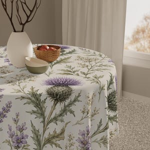 Può includere: Una tovaglia rotonda con una stampa botanica di fiori di cardo viola, foglie verdi e piccoli fiori bianchi su sfondo crema. Il tessuto copre un tavolo circolare su cui sono appoggiati un vaso bianco, una ciotola intrecciata e due ciotole.
