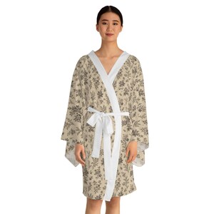 Può includere: Accappatoio beige con stampa floreale, finiture e cintura bianche. L'accappatoio presenta un motivo ripetuto di elementi floreali marrone scuro. La modella indossa l'accappatoio.