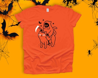 May include: Orange t-shirt featuring a black and white illustration of a cat riding a skeleton figure holding a scythe. The cat has large eyes and the skeleton is wearing a hooded robe.
