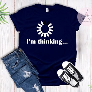 Peut inclure: Un t-shirt bleu marine avec une icône de chargement blanche et le texte "Je réfléchis..." imprimé sur le devant.