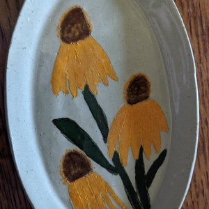 Puede incluir: Un plato ovalado de cerámica blanca con un esmalte moteado presenta un diseño pintado a mano de tres flores amarillas con centros marrones y tallos verdes.