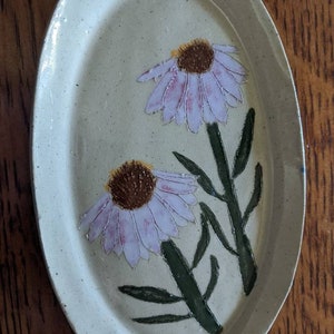 Puede incluir: Un plato ovalado de cerámica blanca con un esmalte moteado presenta dos flores pintadas de morado y rosa con centros marrones y tallos verdes.