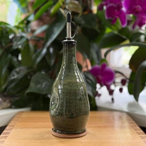 May include: A handcrafted, green ceramic oil bottle with a stainless steel spout. The bottle has a rounded shape and a dark brown base. The background features lush green foliage and vibrant purple orchids, creating a natural aesthetic.