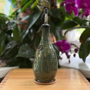 May include: A handcrafted, green ceramic olive oil bottle with a stainless steel spout. The bottle has a textured surface and a decorative emblem. It sits on a wooden base, perfect for kitchen use.