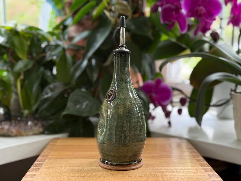 May include: A handcrafted, green ceramic olive oil bottle with a stainless steel spout. The bottle has a textured surface and a decorative emblem. It sits on a wooden base, perfect for kitchen use.
