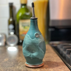 May include: A teal ceramic oil bottle with a stainless steel spout. The bottle has a textured surface and a decorative swirl. The base is a light brown color. Kitchenware.