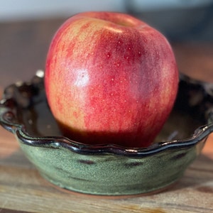 May include: A red apple sits in a green and brown ceramic pie dish with a scalloped rim.