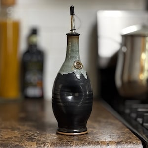 May include: A handcrafted ceramic olive oil dispenser. The bottle is black with a gray-green top and a metal spout. A decorative swirl is on the front. The dispenser is on a countertop in a kitchen setting.