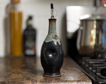May include: A handcrafted ceramic olive oil dispenser. The bottle is black with a gray-green top and a metal spout. A decorative swirl is on the front. The dispenser is on a countertop in a kitchen setting.