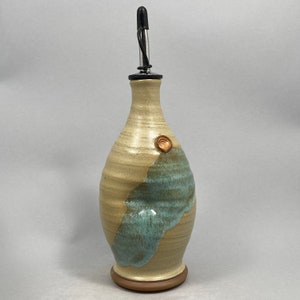 May include: A ceramic olive oil bottle with a light brown and blue glaze. The bottle has a small, round, brown accent on the side. The bottle has a metal pour spout.