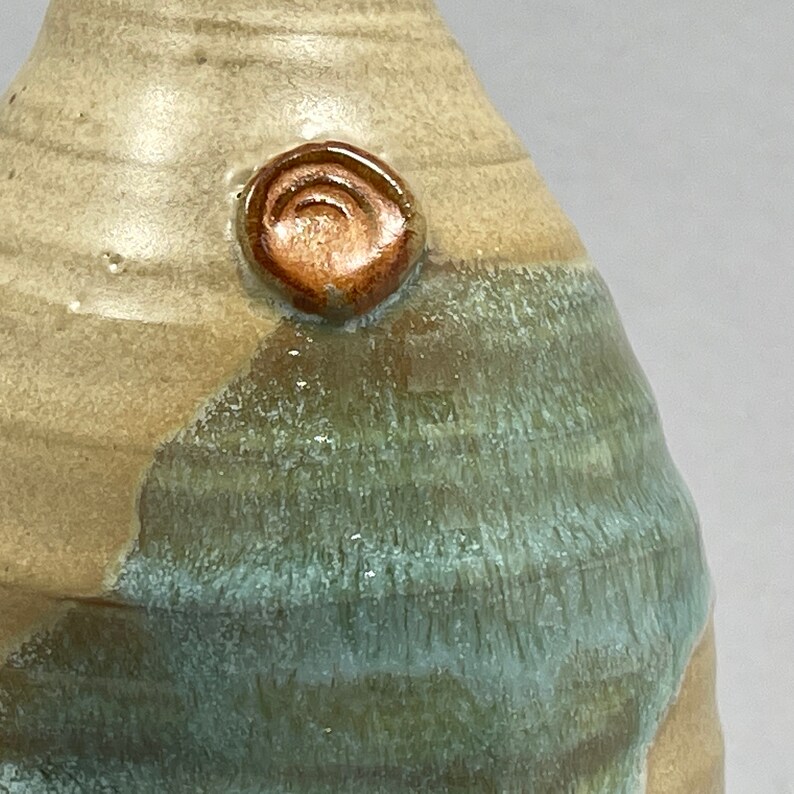 May include: A ceramic vase with a light brown and green glaze. The vase has a round shape and a small, round, brown accent on the side.