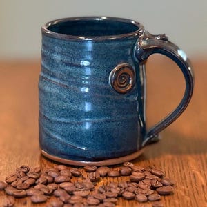 May include: A handmade ceramic mug with a blue glaze and a brown handle. The mug has a spiral design on the side and is surrounded by coffee beans. The mug is approximately 4 inches tall.