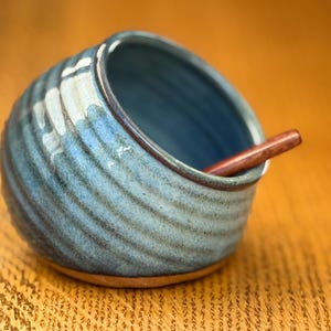 May include: A handmade ceramic salt cellar with a blue and teal glaze. The round bowl has a textured, ridged exterior and a smooth interior. A small wooden spoon rests inside the bowl, ready for use.