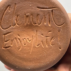 May include: Close-up of a round, brown ceramic object with the words "Clement" and "Enjoy life!" etched into the surface. The text is in a handwritten style. The object has a rough, textured appearance.