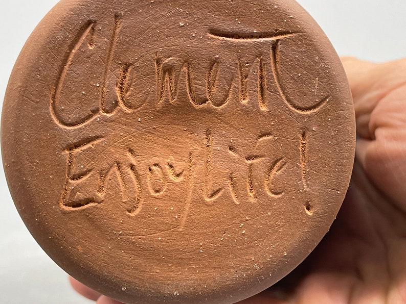 May include: Close-up of a round, brown ceramic object with the words "Clement" and "Enjoy life!" etched into the surface. The text is in a handwritten style. The object has a rough, textured appearance.