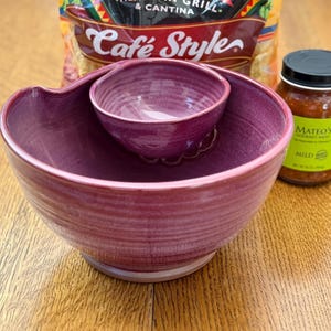 May include: A two-piece, deep purple ceramic chip and dip serving bowl with a smaller bowl nested inside. A jar of Mateo's Gourmet Salsa and a bag of Cafe Style tortilla chips are in the background. The bowl is on a wooden surface.