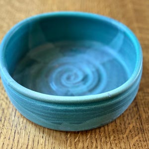 May include: A handmade ceramic bowl in shades of teal and turquoise. The bowl has a rounded shape with a slightly flared rim. The interior features a spiral design. This decorative bowl is suitable for serving or display.