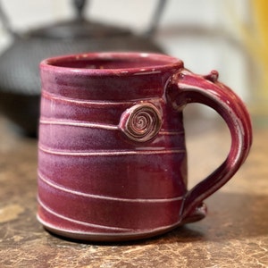 May include: A handmade ceramic mug with a deep burgundy glaze and white stripes. The mug has a unique, slightly asymmetrical handle and a small spiral design on the front.