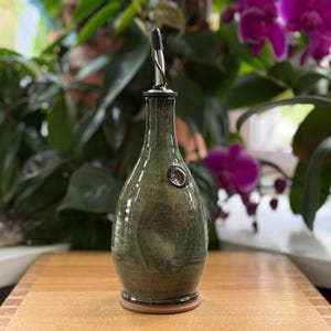 May include: A handcrafted olive oil bottle with a dark green glaze. The bottle has a long neck, a metal spout, and a decorative seal. It sits on a wooden surface, with a wooden base. The background features lush green plants and vibrant purple orchids.