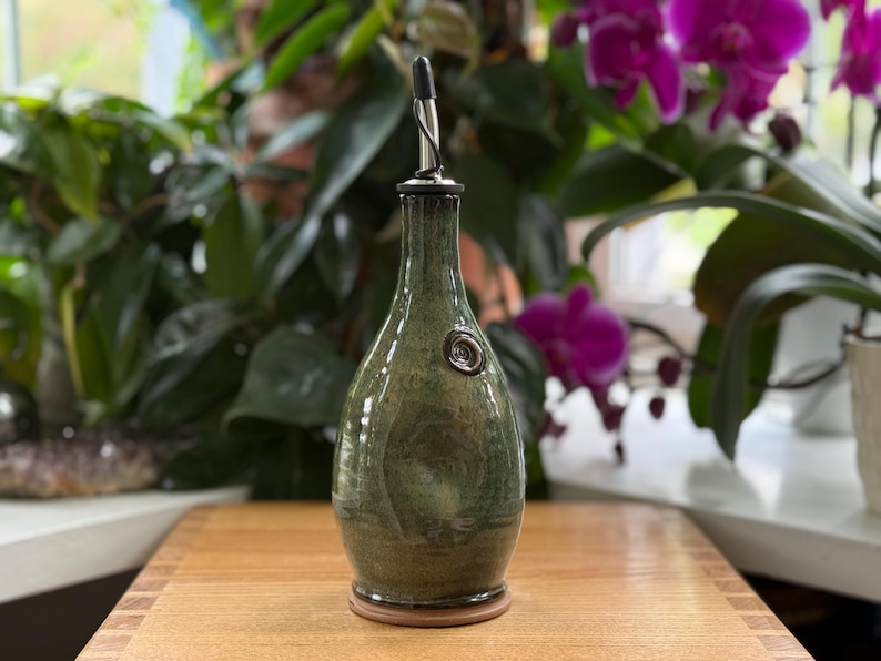 May include: A handcrafted olive oil bottle with a dark green glaze. The bottle has a long neck, a metal spout, and a decorative seal. It sits on a wooden surface, with a wooden base. The background features lush green plants and vibrant purple orchids.
