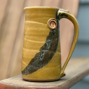 May include: A handmade ceramic mug with a large handle. The mug is a golden-yellow color with a dark brown and green diagonal stripe. A small spiral design is on the side of the mug. The mug is sitting on a wooden surface.