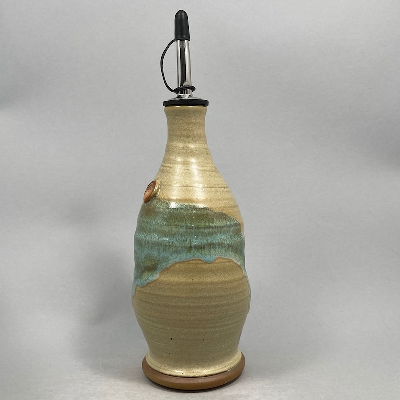 May include: A ceramic olive oil bottle with a light brown and blue glaze. The bottle has a metal pour spout and a small brown knob on the side.