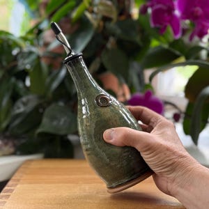 May include: A handcrafted, green ceramic oil bottle with a stainless steel spout. The bottle has a unique, organic shape and a decorative metal accent. The bottle is held in a hand, showcasing its size and design. The background features lush green plants and purple flowers.
