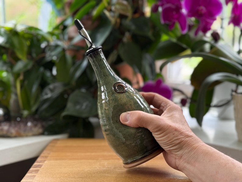 May include: A handcrafted, green ceramic oil bottle with a stainless steel spout. The bottle has a unique, organic shape and a decorative metal accent. The bottle is held in a hand, showcasing its size and design. The background features lush green plants and purple flowers.