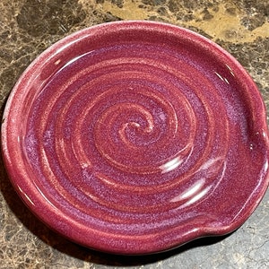 May include: A handmade, ceramic spoon rest in a deep burgundy color with a glossy finish. The circular rest features a spiral design in varying shades of red and purple. The spoon rest has a small, curved indentation on one side.