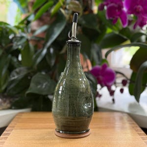 May include: A green ceramic olive oil bottle with a stainless steel spout. The bottle has a bulbous shape and a dark brown base. The background features lush green plants and purple orchids, set on a wooden surface.