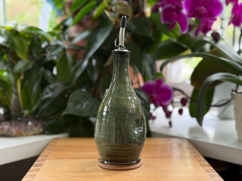 May include: A green ceramic olive oil bottle with a stainless steel spout. The bottle has a bulbous shape and a dark brown base. The background features lush green plants and purple orchids, set on a wooden surface.