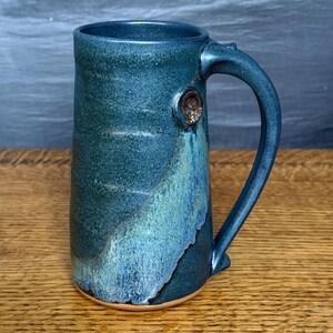 May include: A tall, handcrafted ceramic mug with a large handle. The mug features a dark teal glaze with lighter blue and tan accents. A small, decorative element is affixed to the side of the mug near the handle. The mug sits on a wooden surface.