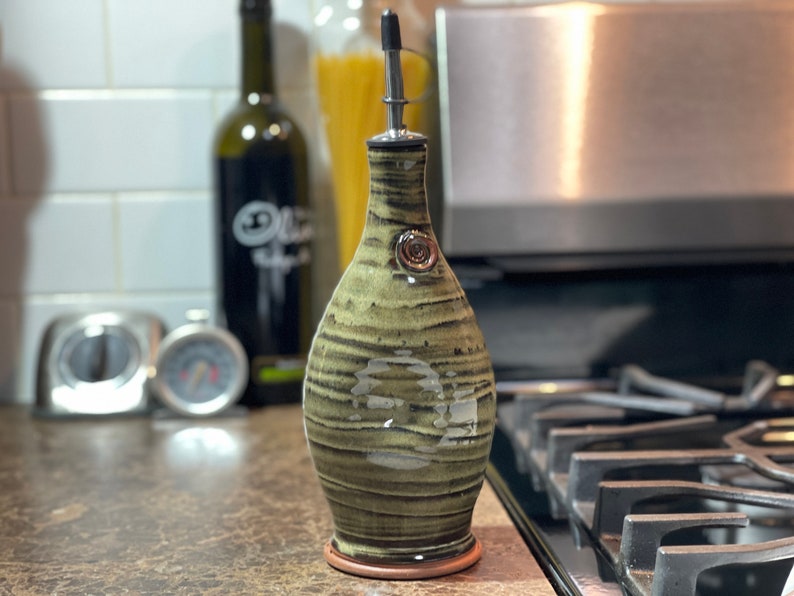May include: A handcrafted olive oil dispenser with a unique, earthy design. The ceramic bottle features a green and brown striped pattern, a metal spout, and a decorative metal accent. The dispenser is approximately 8 inches tall.