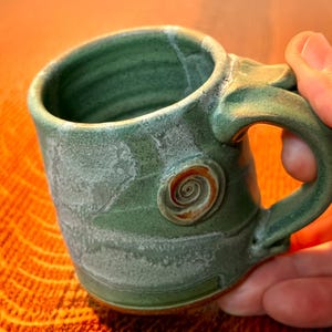 May include: Handmade ceramic mug in shades of green with a spiral design. The mug has a rounded shape and a sturdy handle. The mug is approximately 3 inches tall and 4 inches wide.