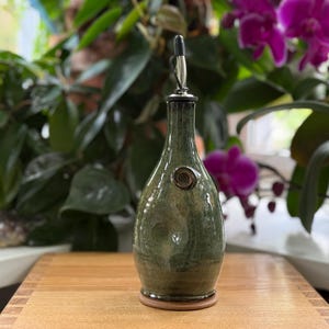 May include: A green ceramic oil bottle with a metal spout and a decorative swirl. The bottle has a dark green glaze with lighter green and brown accents. The bottle is on a wooden surface.