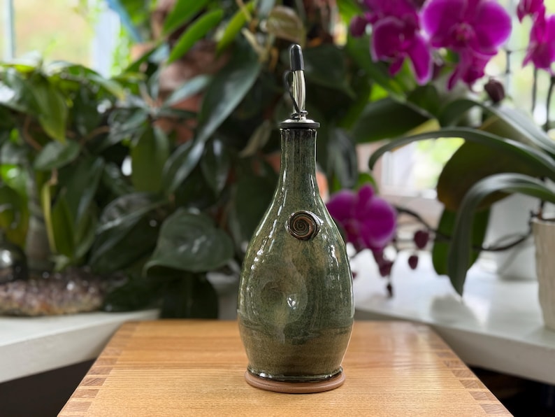 May include: A green ceramic oil bottle with a metal spout and a decorative swirl. The bottle has a dark green glaze with lighter green and brown accents. The bottle is on a wooden surface.