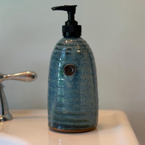 May include: A blue ceramic soap dispenser with a black pump. The dispenser has a textured surface and a small, decorative brown accent. The dispenser is on a white surface, with a chrome faucet visible in the background.
