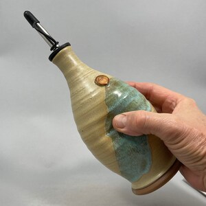 May include: A ceramic olive oil bottle with a light brown and blue glaze. The bottle has a spout with a stainless steel pourer and a cork stopper. The bottle is hand-thrown and has a unique, rustic look.
