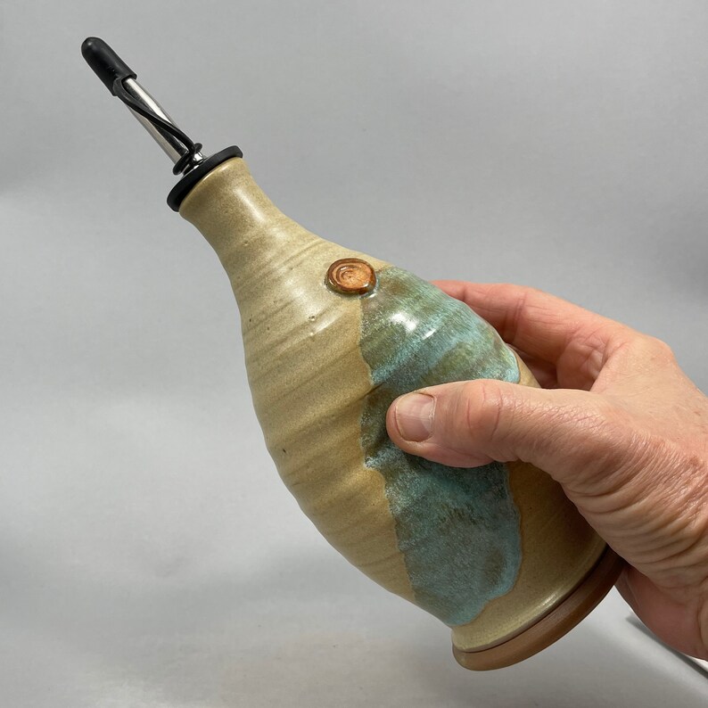 May include: A ceramic olive oil bottle with a light brown and blue glaze. The bottle has a spout with a stainless steel pourer and a cork stopper. The bottle is hand-thrown and has a unique, rustic look.